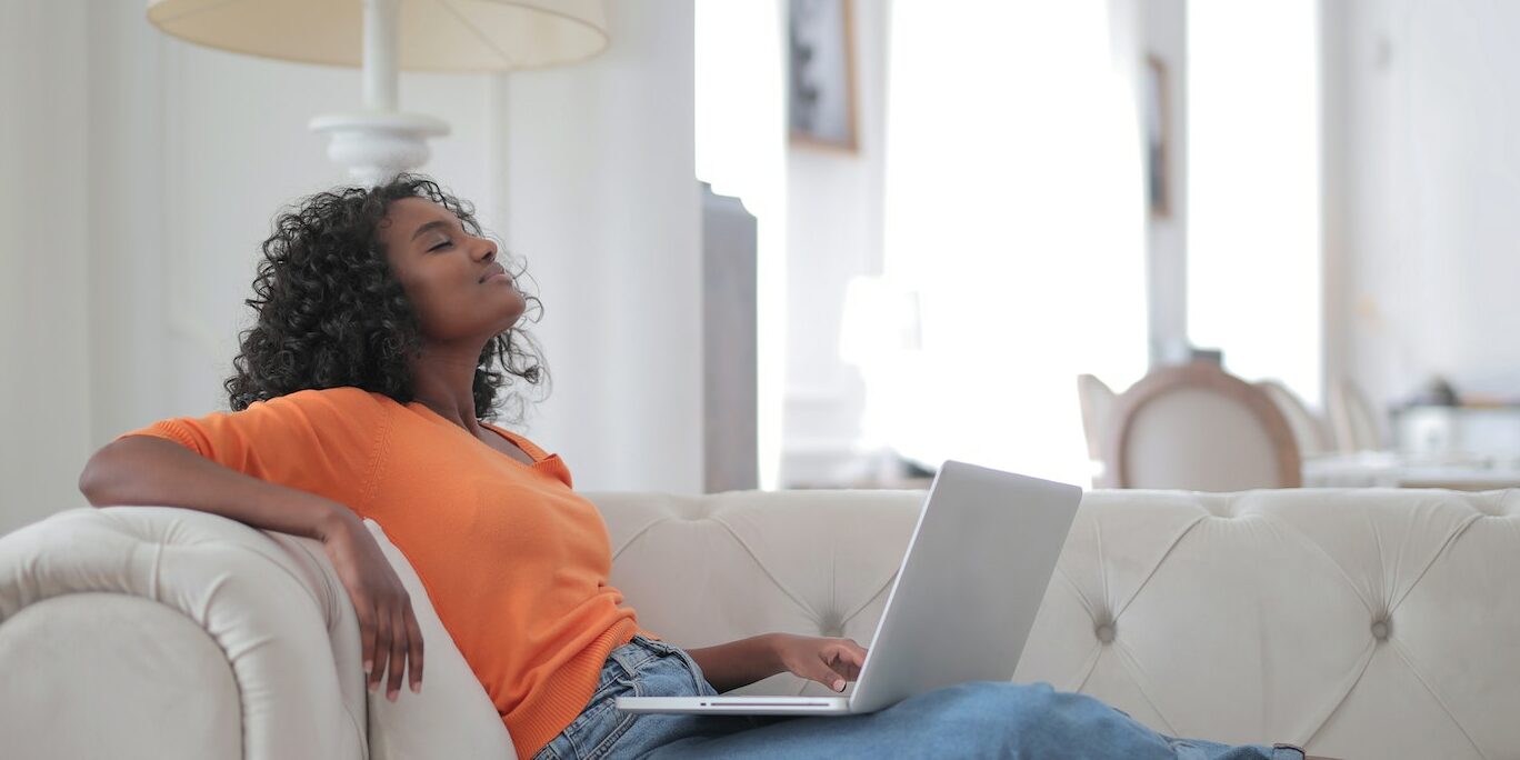 Woman in Orange Top and Blue Denim Jeans Sitting on White Sofa Using Macbook